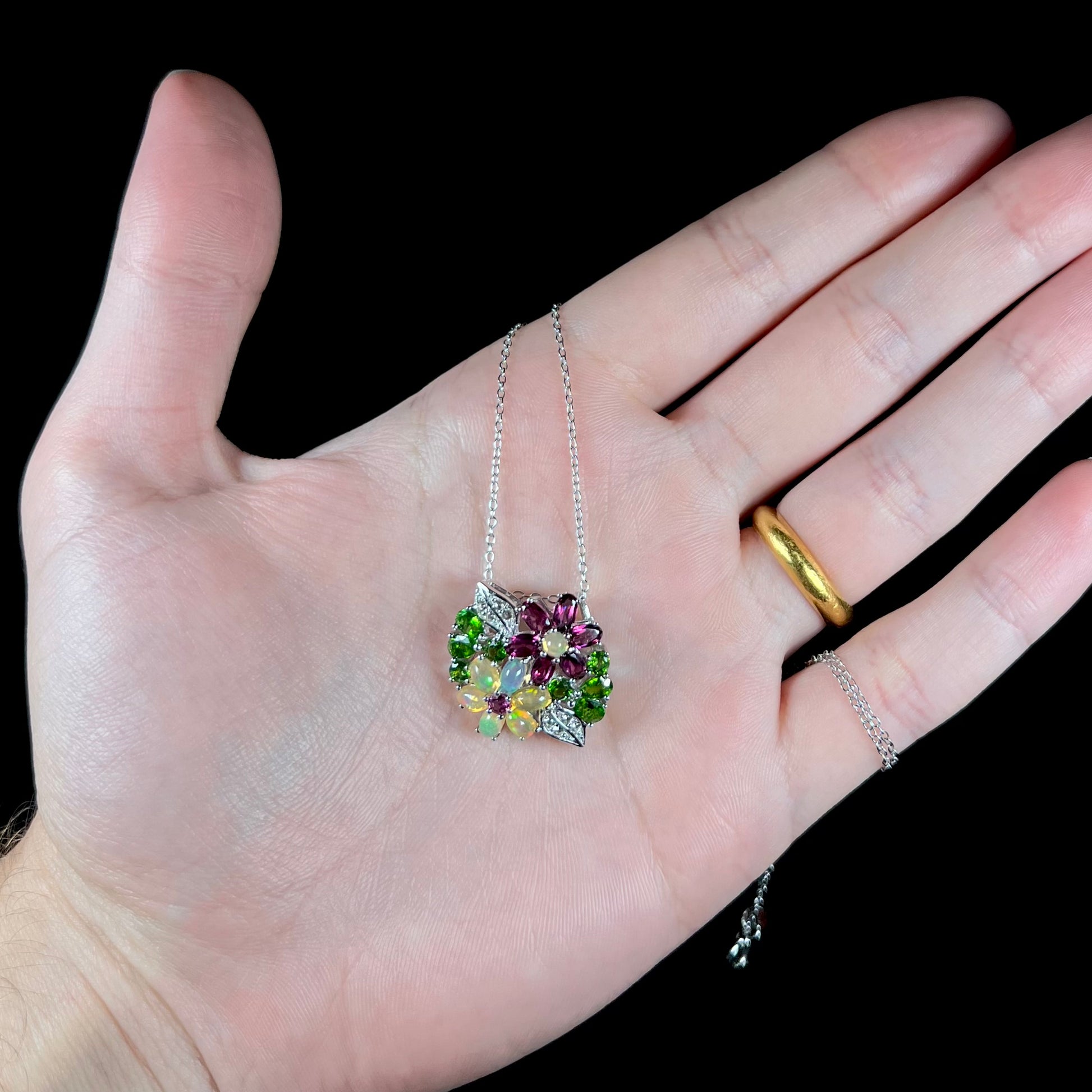 An opal, garnet, and chrome diopside flower necklace held in a hand against a black background.