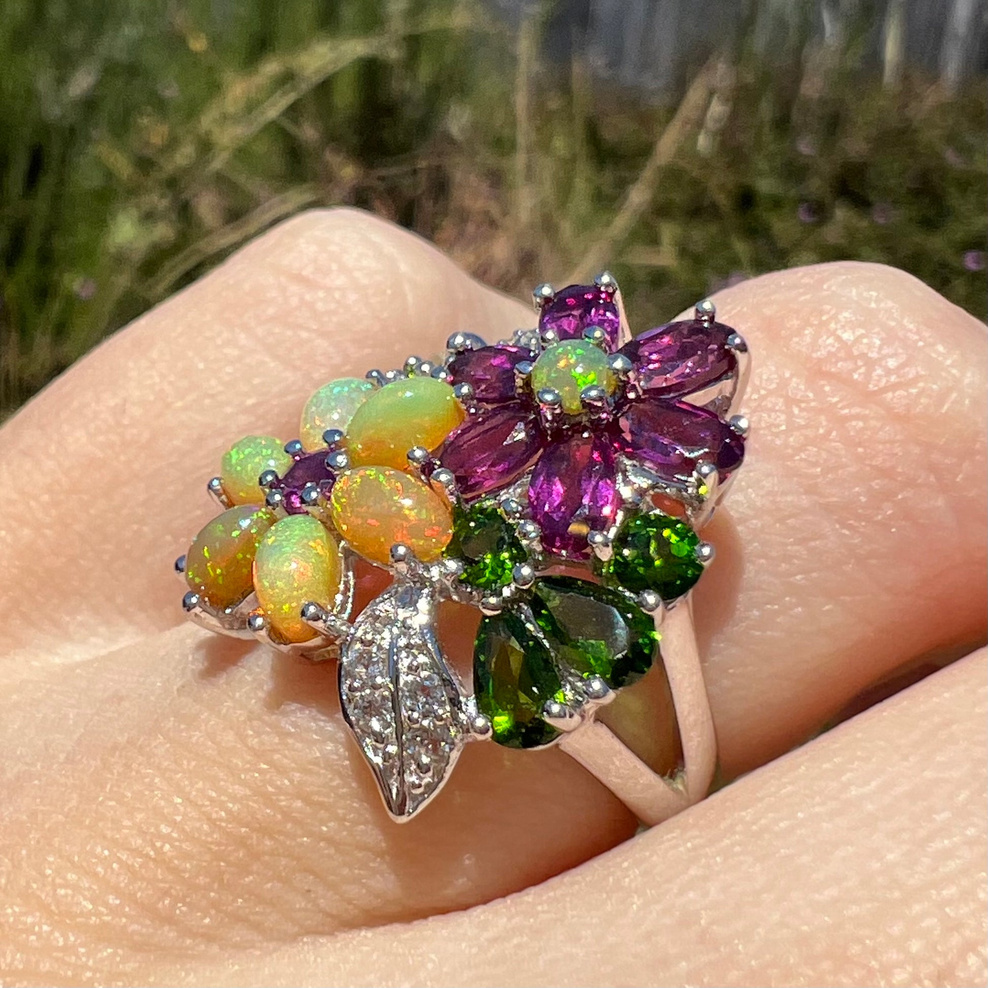 An opal and garnet gemstone flower ring, shown at an angle on a finger close-up in the sunlight.