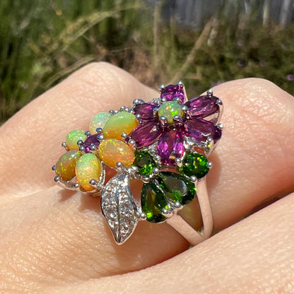 An opal and garnet gemstone flower ring, shown at an angle on a finger close-up in the sunlight.
