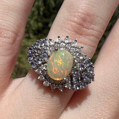 Close-up sunlit view of Ethiopian opal ring with spray of tanzanites and white topaz, worn on finger