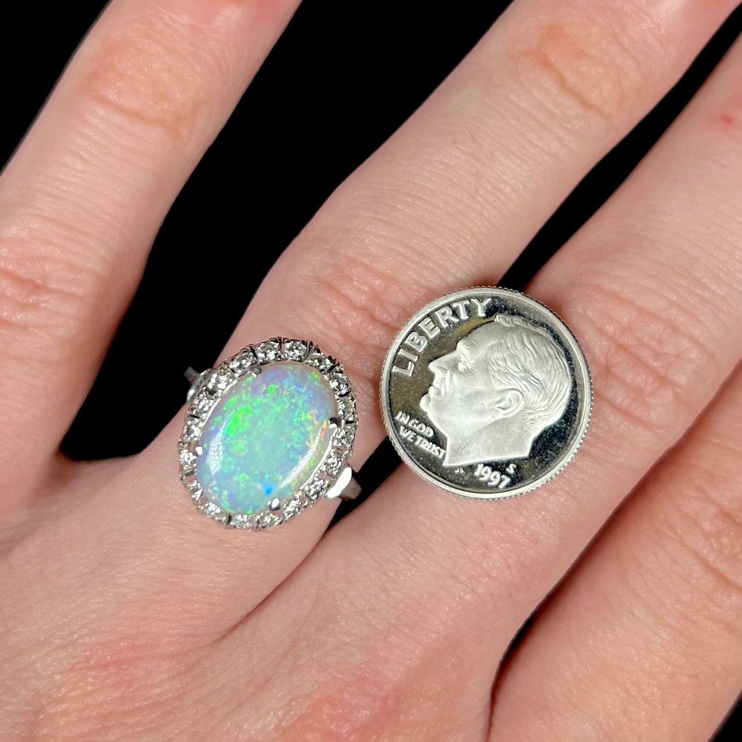 An opal and diamond halo ring on a hand next to a US dime coin for scale.