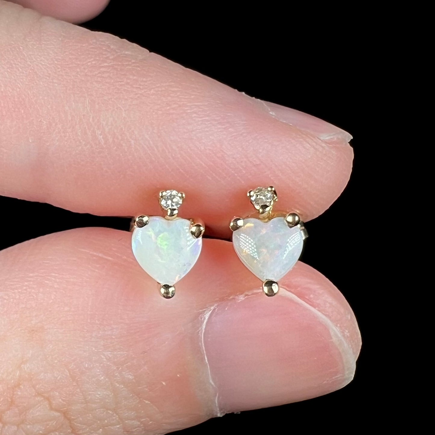 Close-up of 14k gold opal heart stud earrings with diamond accents, held between two fingers.