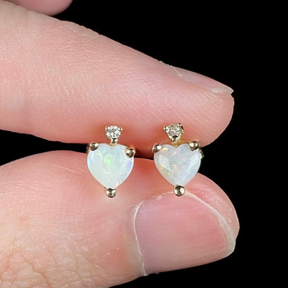 Close-up of 14k gold opal heart stud earrings with diamond accents, held between two fingers.