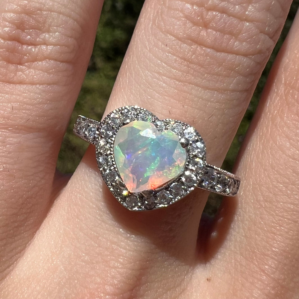Close-up of faceted heart shaped Ethiopian opal ring with CZ halo design, worn on finger in sunlight