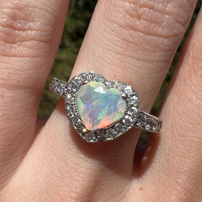 Close-up of faceted heart shaped Ethiopian opal ring with CZ halo design, worn on finger in sunlight