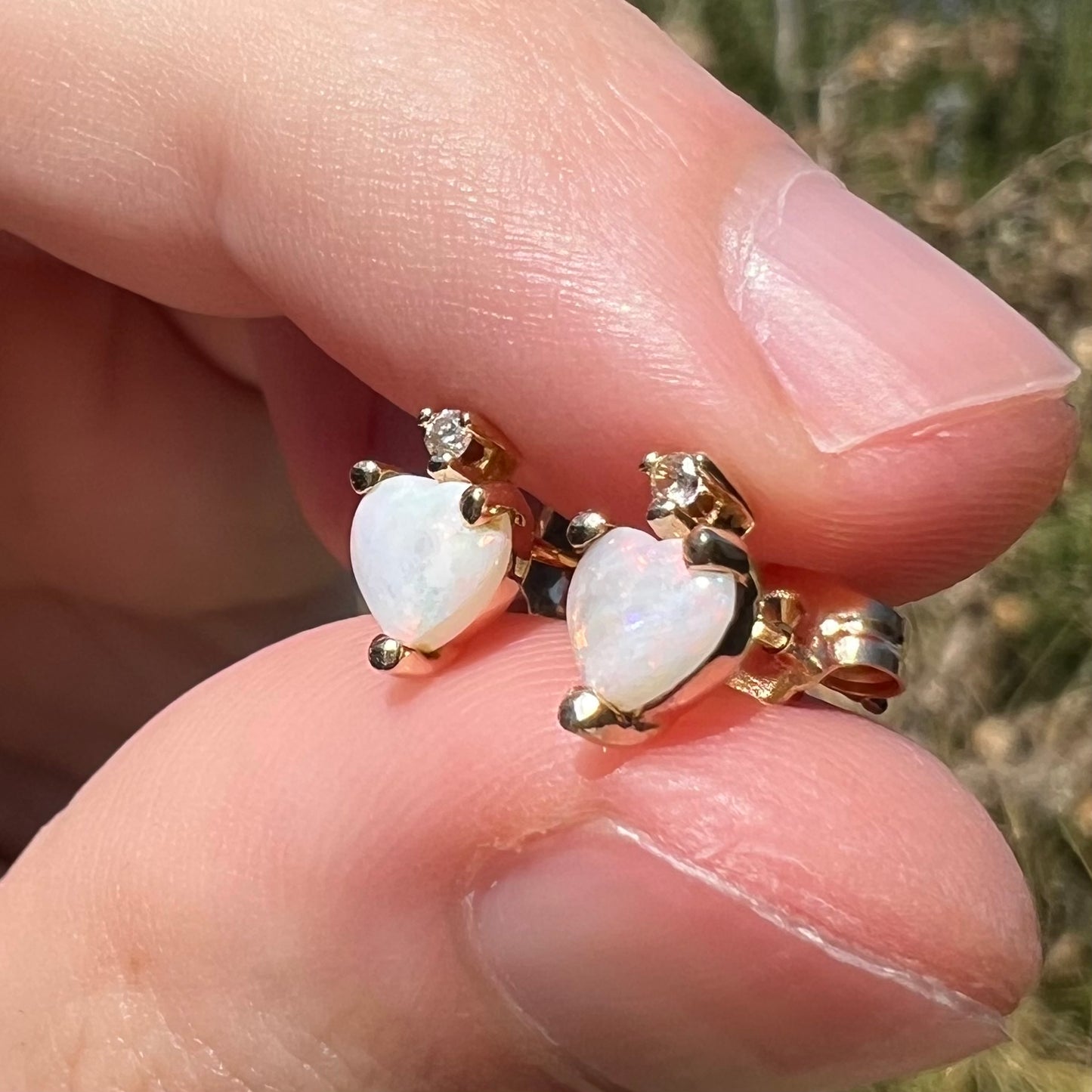 Heart shaped white Australian opal stud earrings in 14k yellow gold with diamond accents, shown close-up from angle in sunlight held between two fingers.