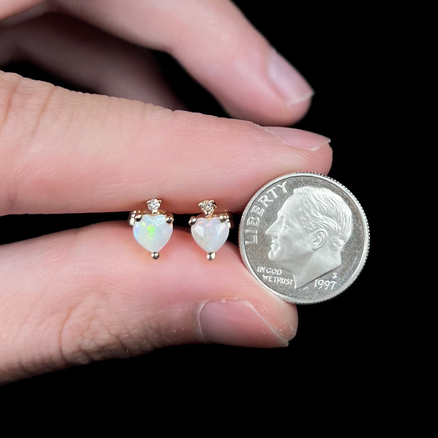 Fingers holding heart shaped opal stud earrings in gold next to US dime coin for scale.