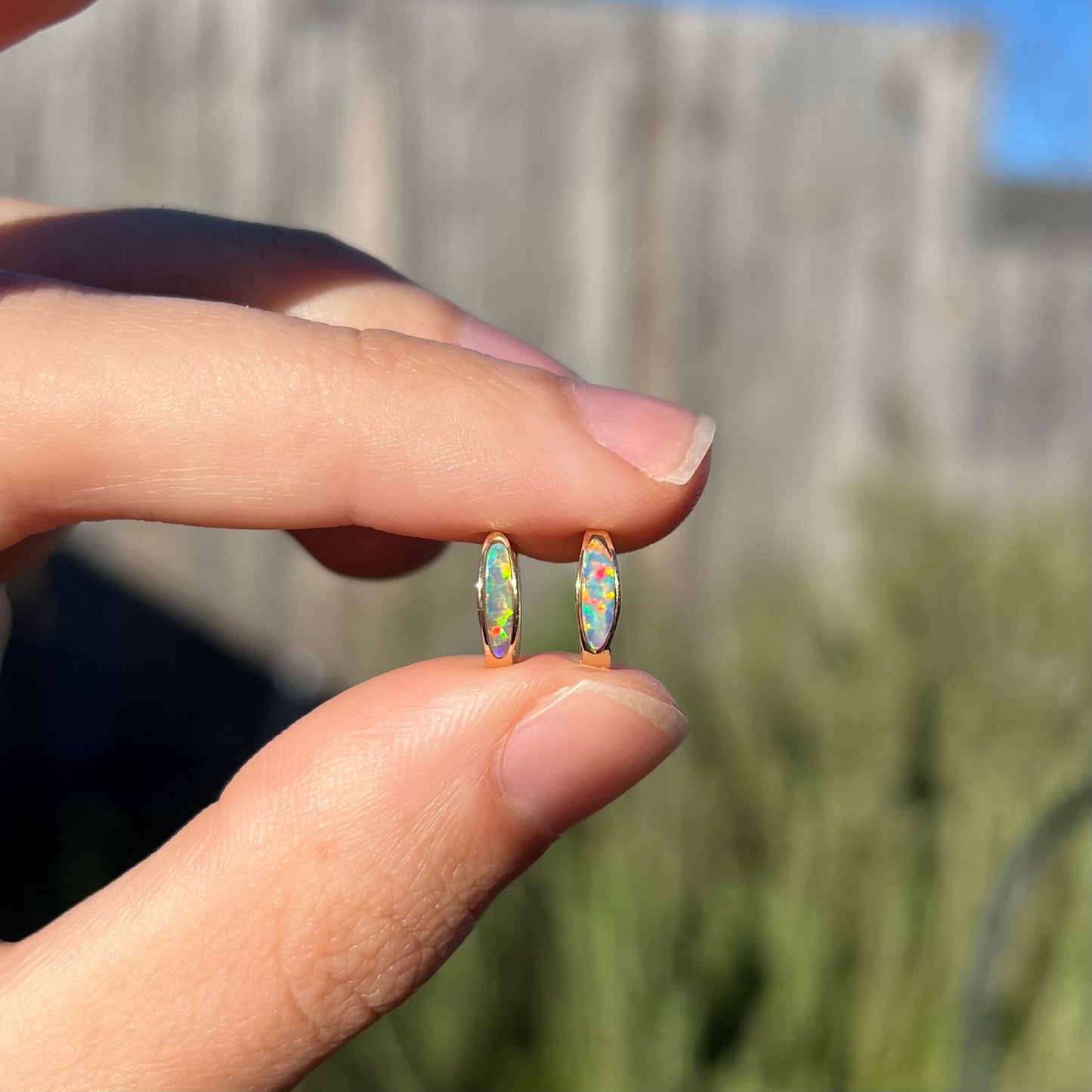 A pair of yellow gold lab-created opal inlay huggie hoop earrings held between two fingers in the sunlight.