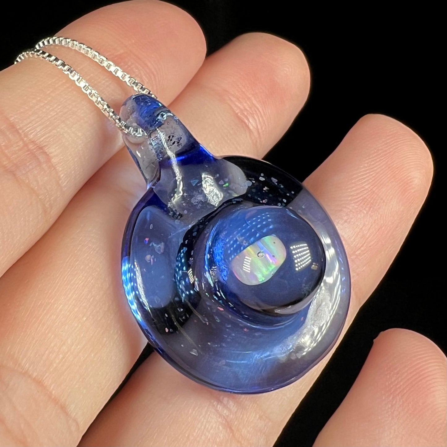 Anlged close-up of a lab-created opal in  a domed blue glass pendant fitted to a silver chain, held in a hand.