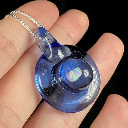Anlged close-up of a lab-created opal in  a domed blue glass pendant fitted to a silver chain, held in a hand.