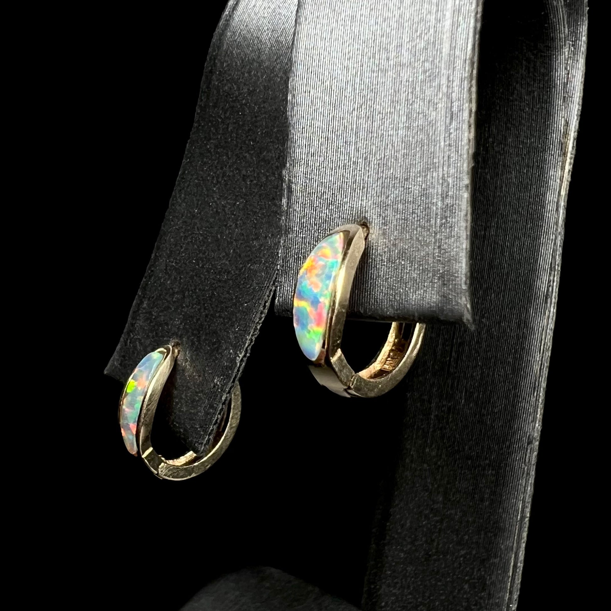 Angled view of a pair of yellow gold lab-created opal inlay huggie hoop earrings on an earring display against a black background.