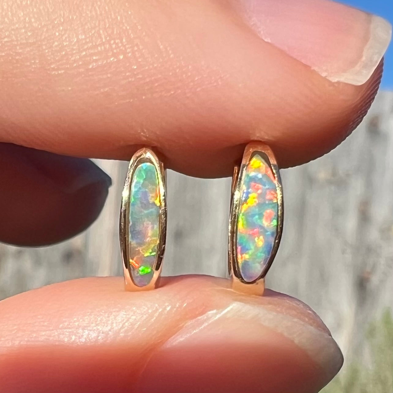 Close-up of a pair of yellow gold lab-created opal inlay huggie hoop earrings held between two fingers in sunlight.