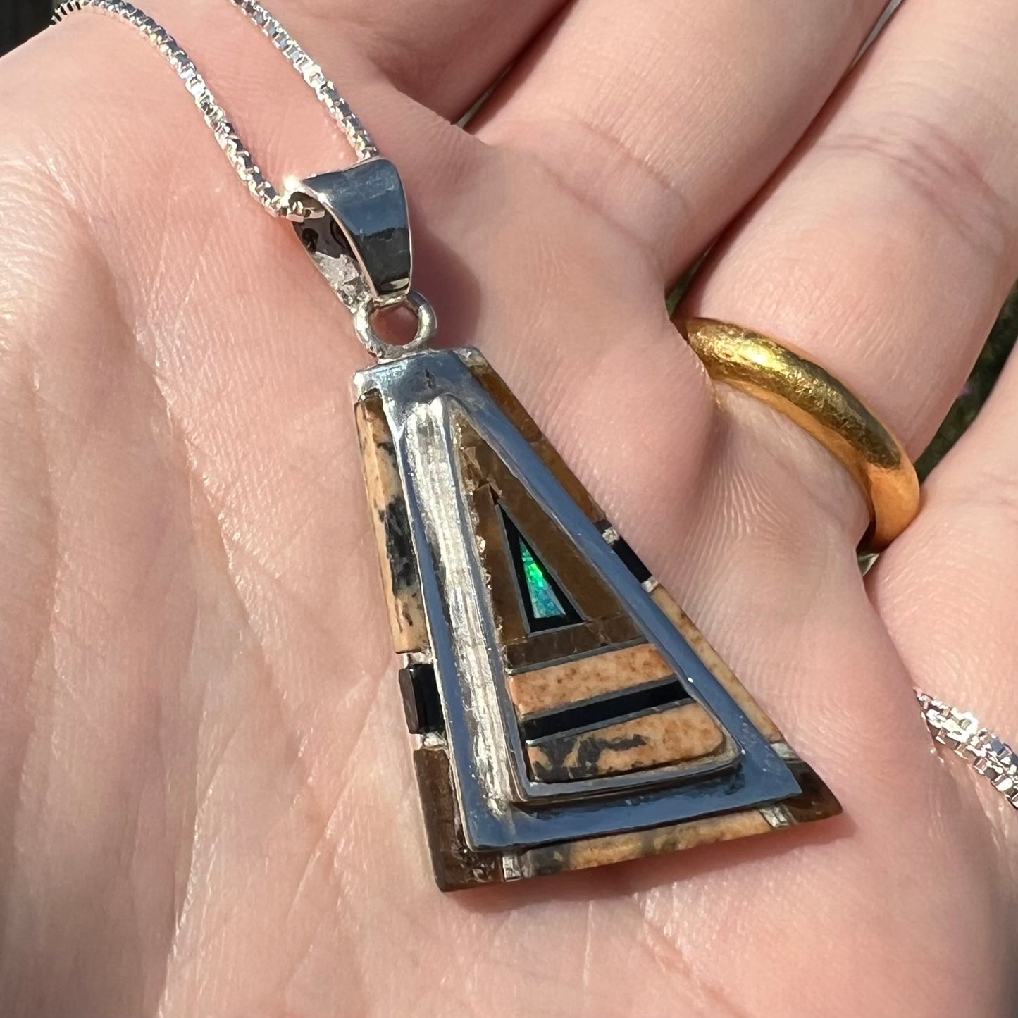 A sterling silver Australian opal, picture jasper, and onyx inlay pendant shown close-up at an angle in sunlight.