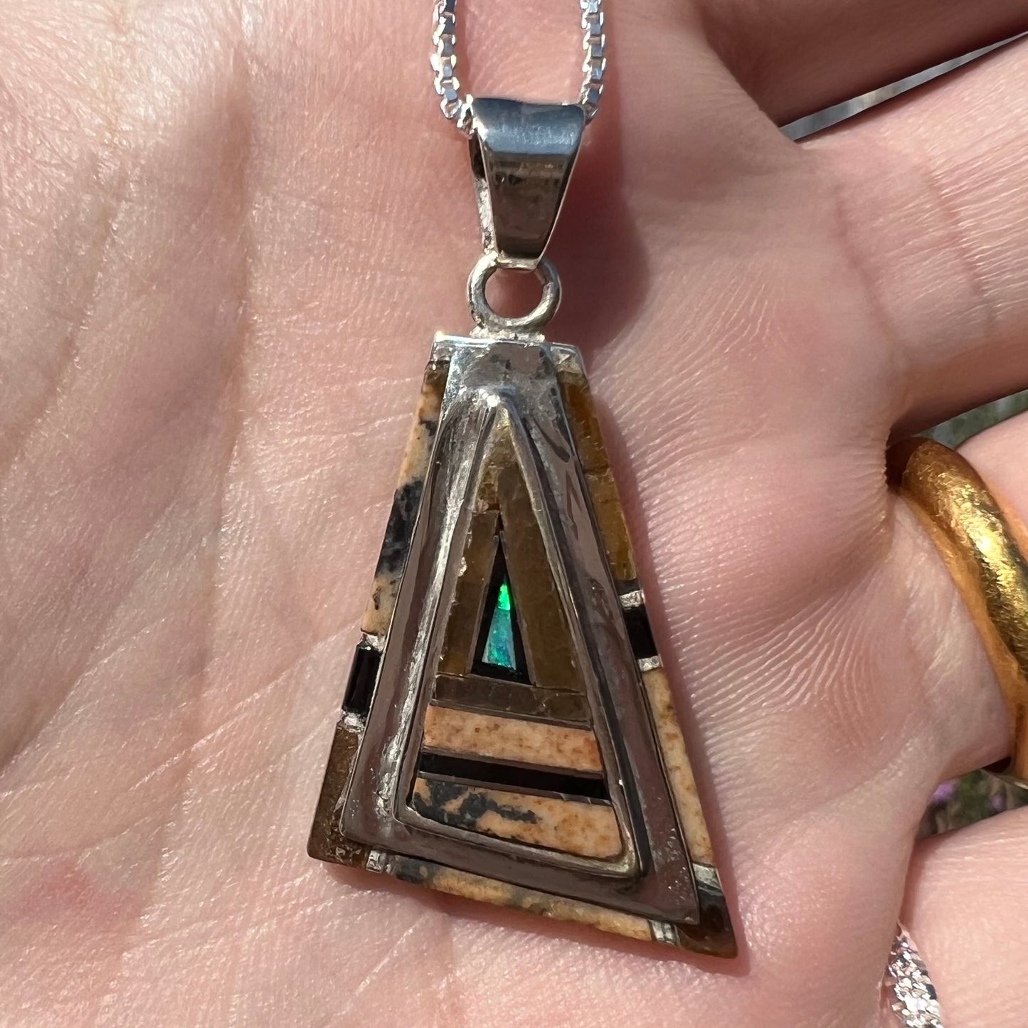 Close-up of a sterling silver Australian opal, picture jasper, and onyx inlay pendant in sunlight, held in a hand.
