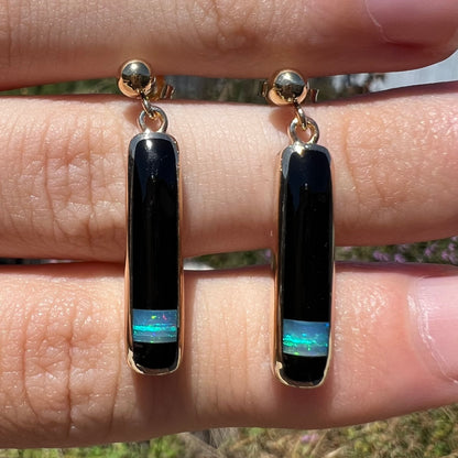 Close-up of opal and onyx inlay dangle earrings shown from the front view, held in the sunlight.