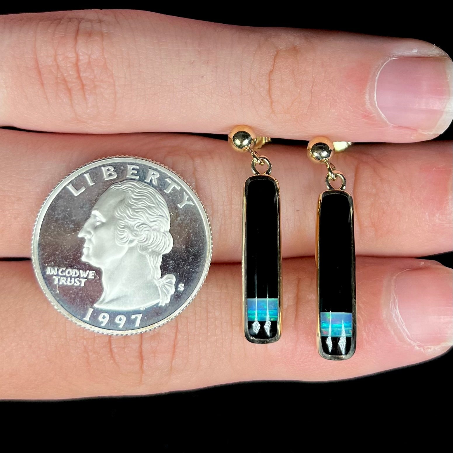 A pair of opal and onyx dangle earrings held between fingers next to a 1997 S quarter coin for scale.