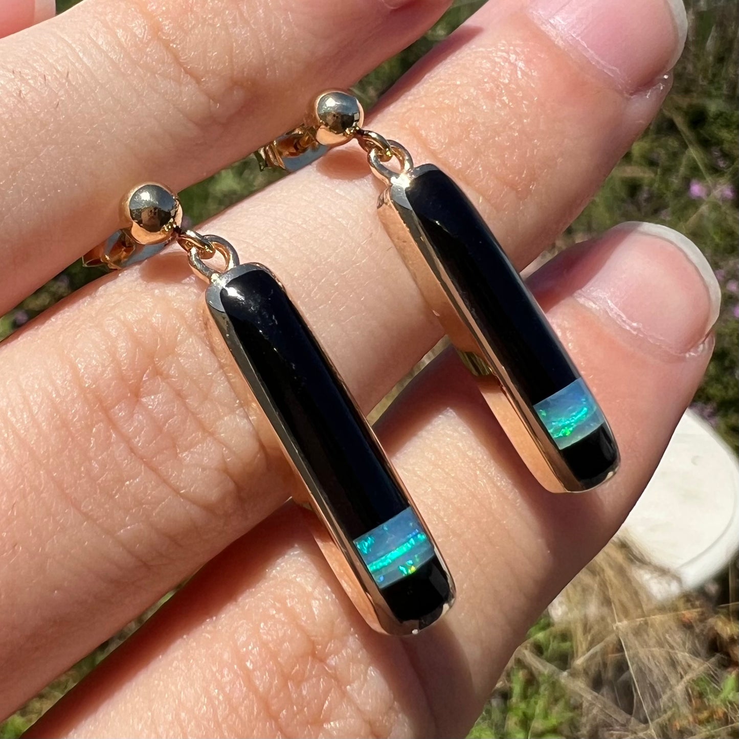A pair of opal and onyx inlay drop earrings shown close-up at an angle, held between fingers in natural light.