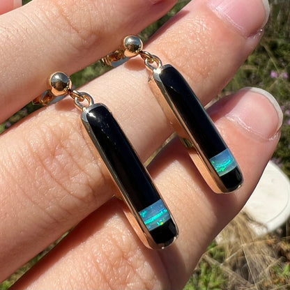 A pair of opal and onyx inlay drop earrings shown close-up at an angle, held between fingers in natural light.