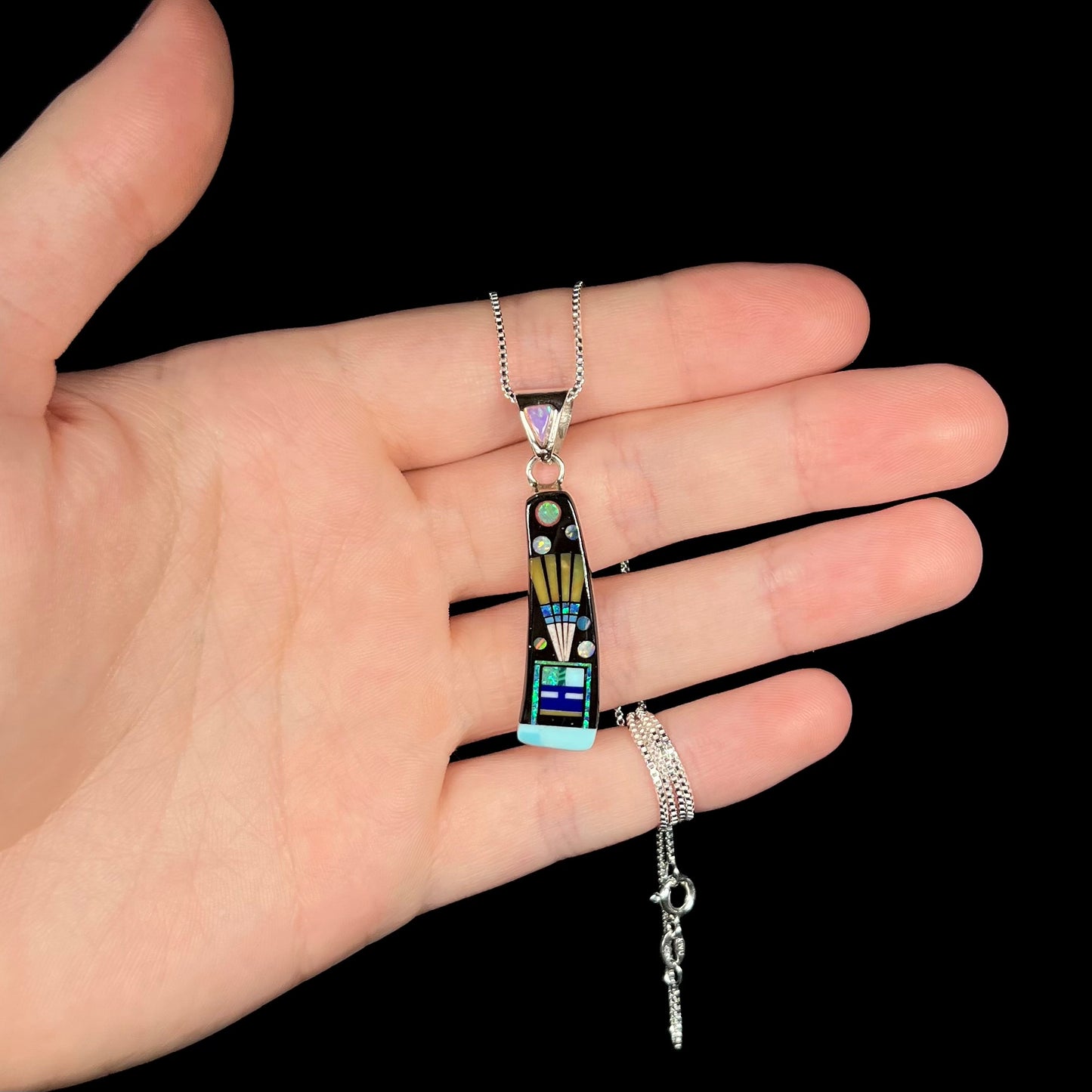 Hand holding an opal and onyx gemstone inlay necklace with Southwest-style kachina motif against black background.