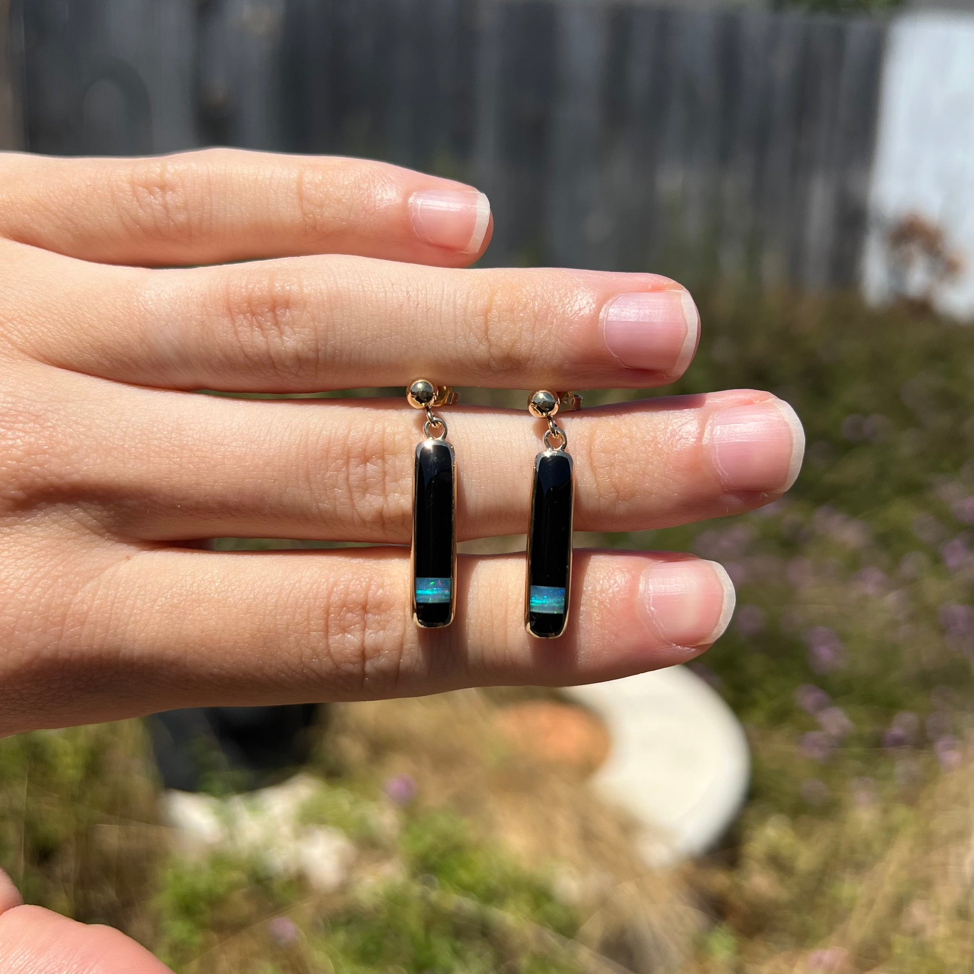 A pair of dangling opal and onyx inlay earrings held between fingers at a distance in the sunlight.