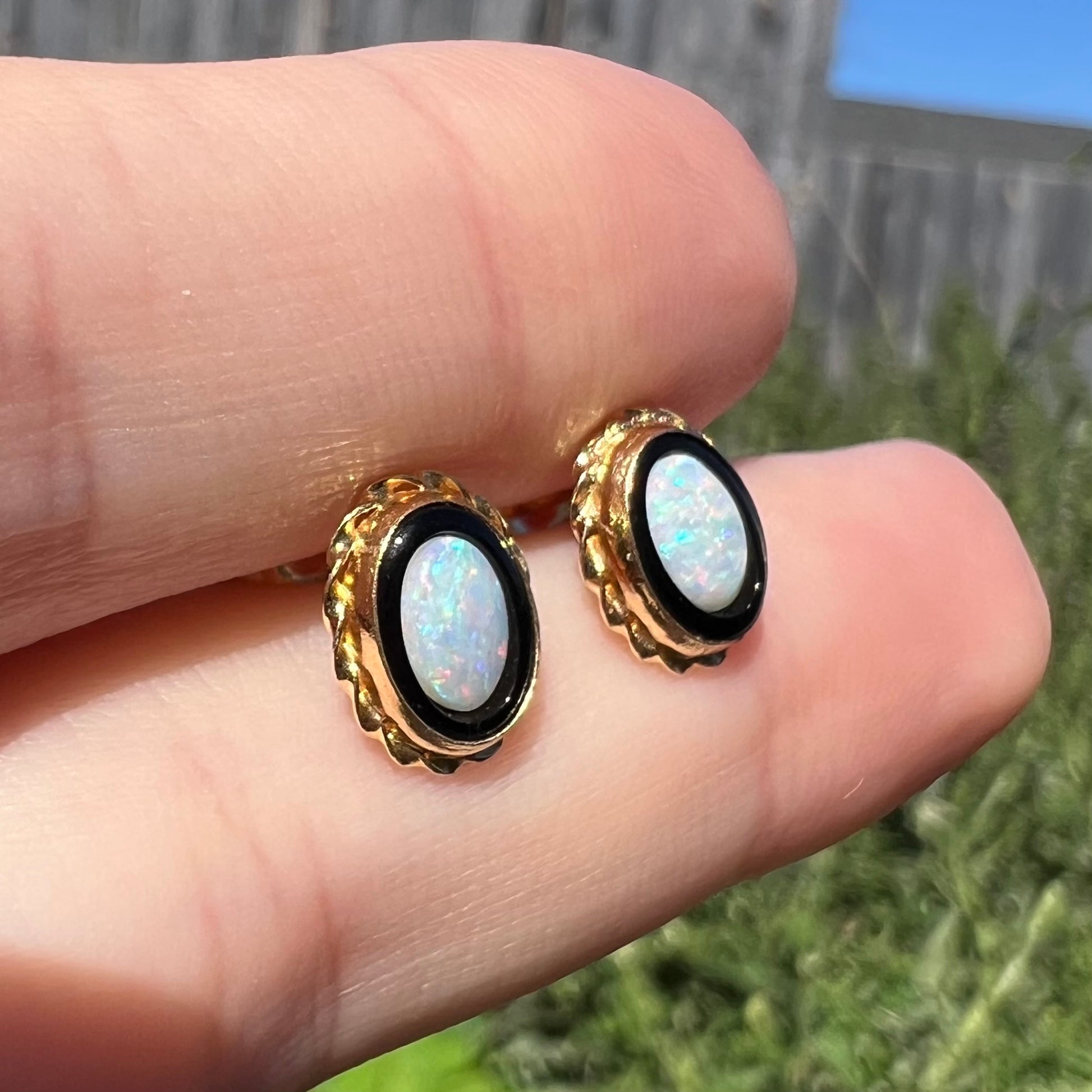 Angled close-up of opal and onyx stud earrings with twisted gold rope design held between two fingers in natural light against outdoor backdrop.