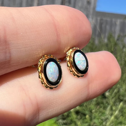 Angled close-up of opal and onyx stud earrings with twisted gold rope design held between two fingers in natural light against outdoor backdrop.
