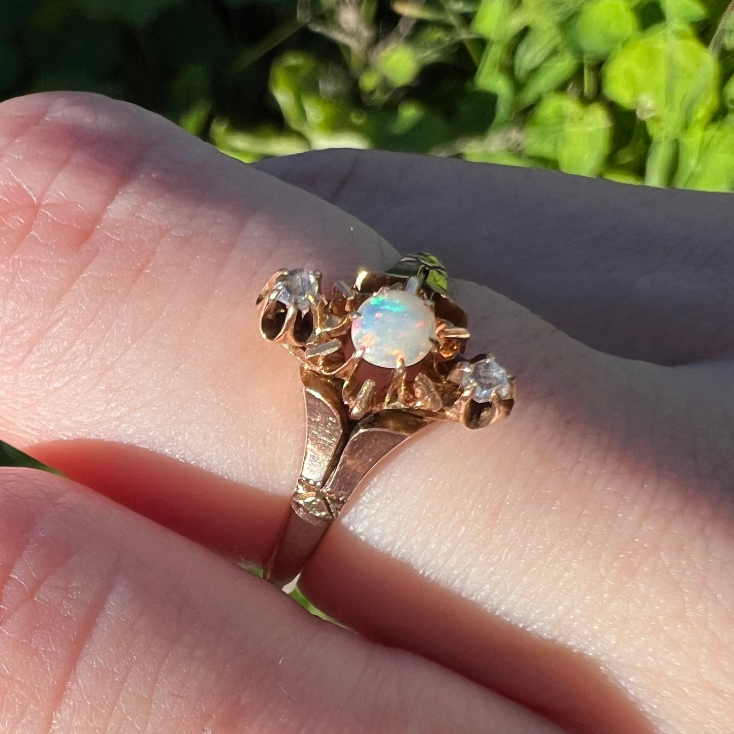 An English gold opal and rose cut diamond ring shown close-up from an angle on a finger in natural light against a blurred green backdrop.