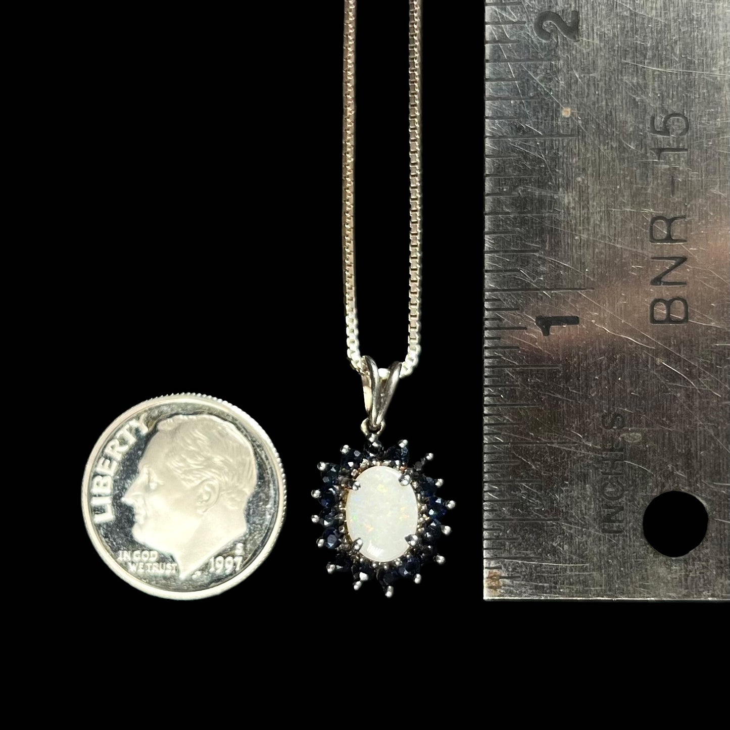 A silver opal and sapphire halo necklace next to a dime and ruler for size comparison, measuring approximately 0.75 inches including the bail.