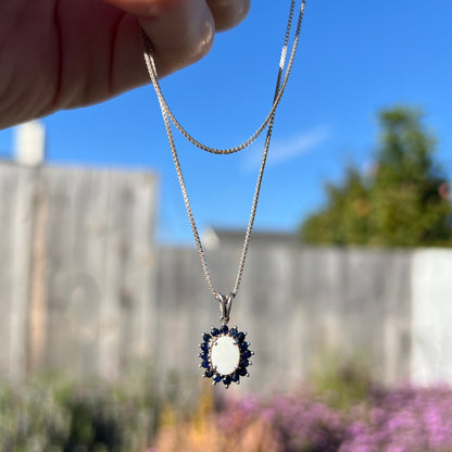 A sterling silver opal and sapphire necklace being dangled by its chain outdoors in natural sunlight.