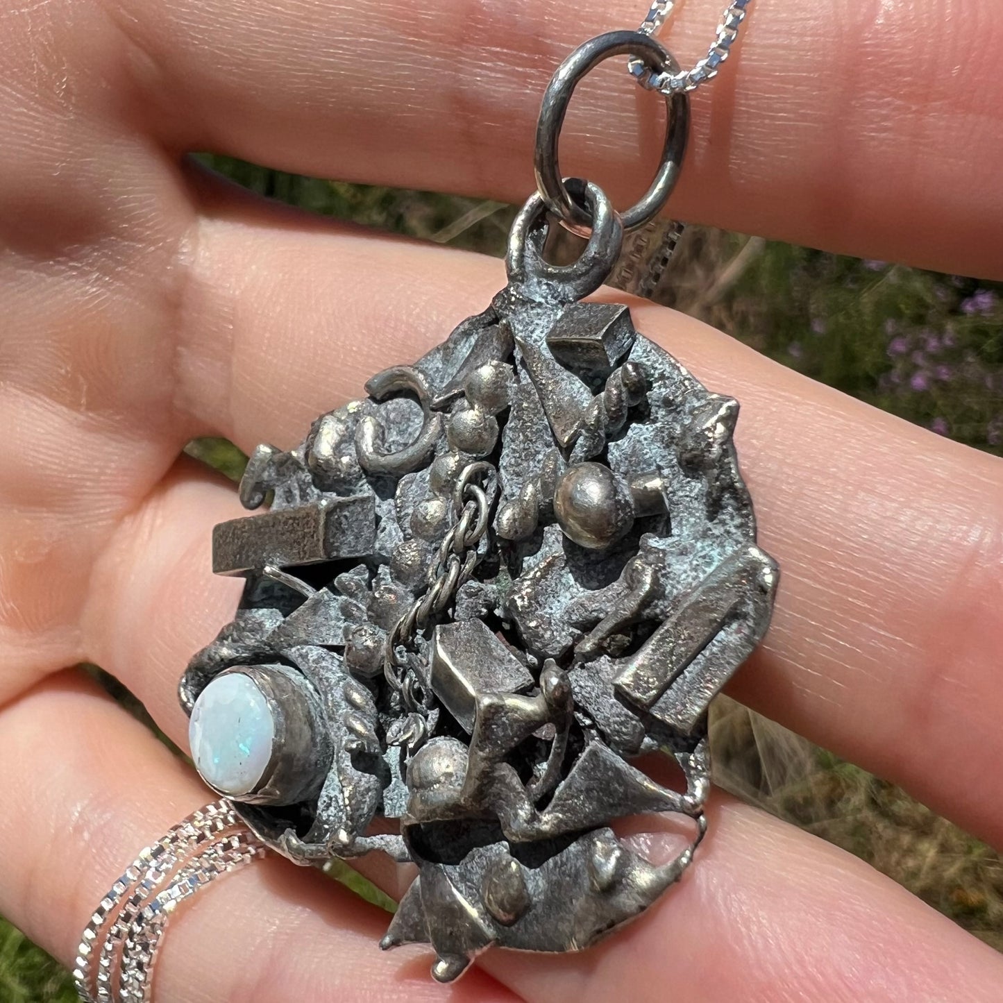 A brutalist-style opal shipwreck pendant made from silver, shown at an angle close-up in the sunlight.