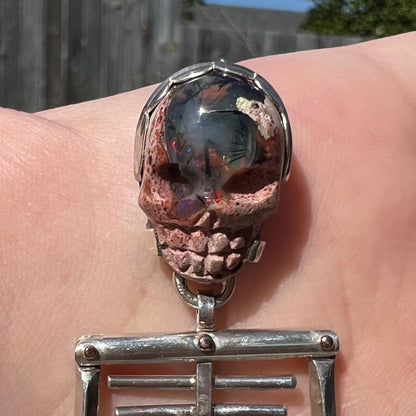 Close-up of carved Mexican cantera opal skull, mounted in a two-tone sterling silver and 14k gold skeleton pendant, held in sunlight