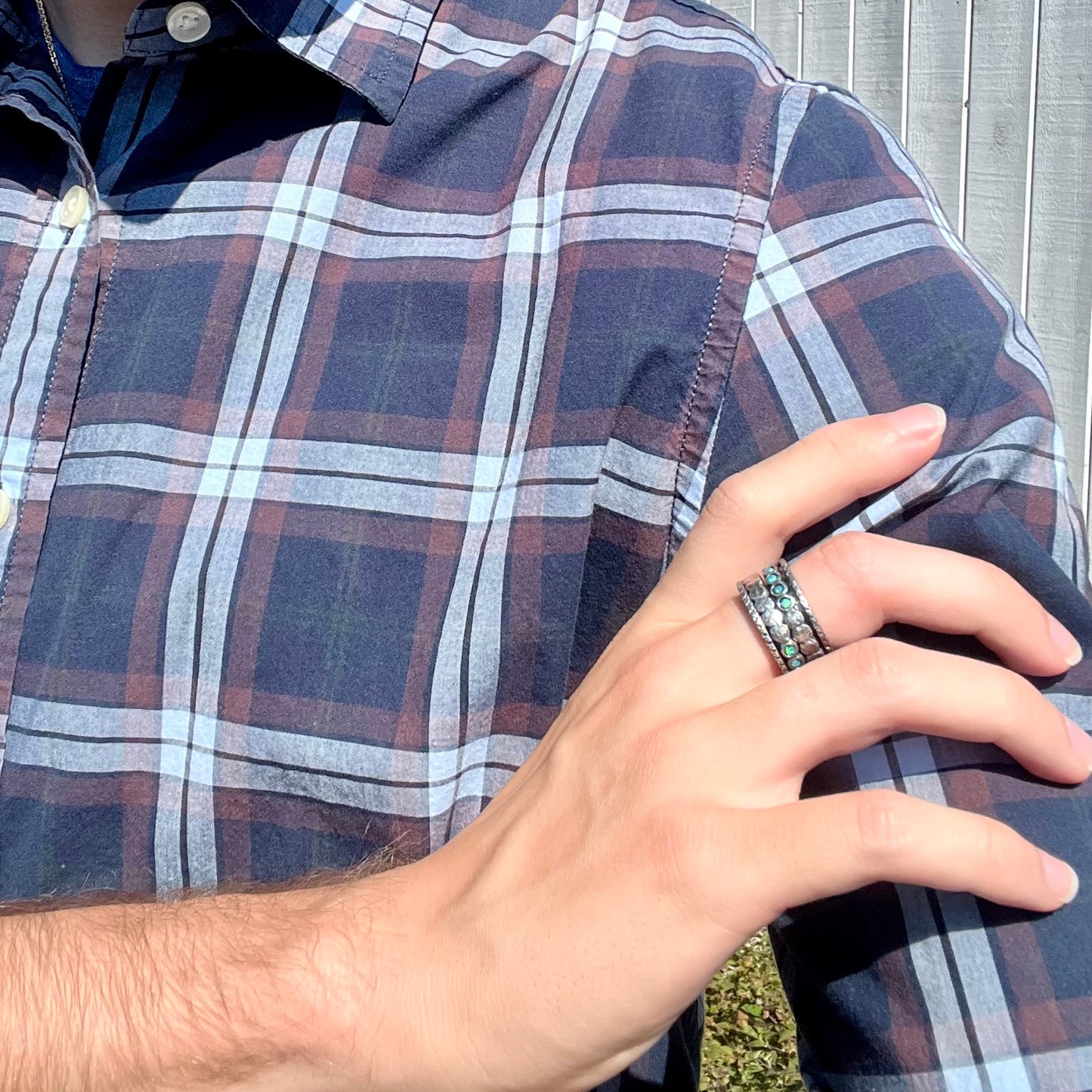 Person in plaid shirt crossing arm to hold opposing shoulder while wearing a men's opal spinner ring.