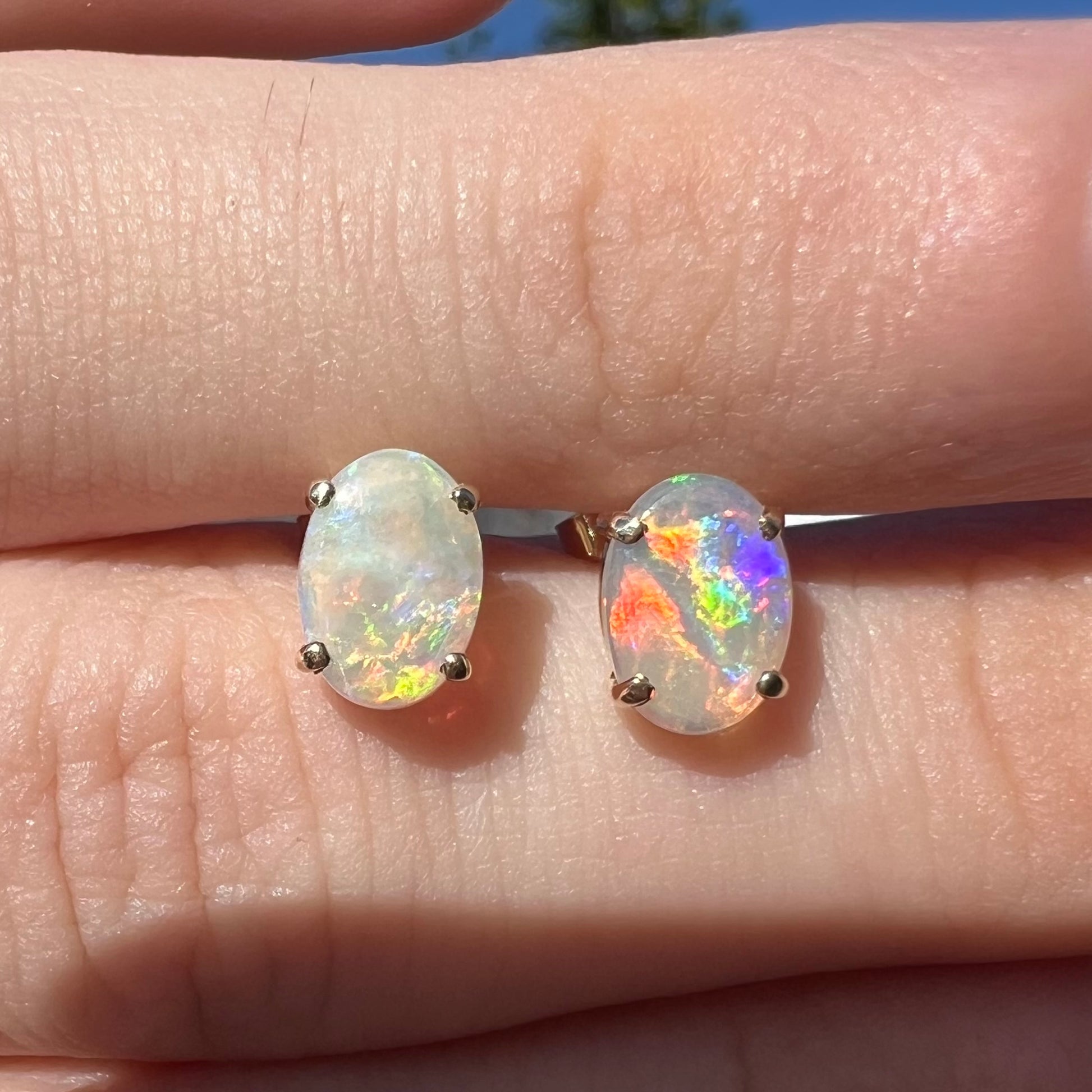 A macro close-up of opal stud earrings in natural sunlight, one opal displaying vibrant multicolor flashes while the other has faint yellow flashes.