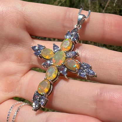 A silver opal and tanzanite cross necklace, held in the sunlight by a hand at an angle.