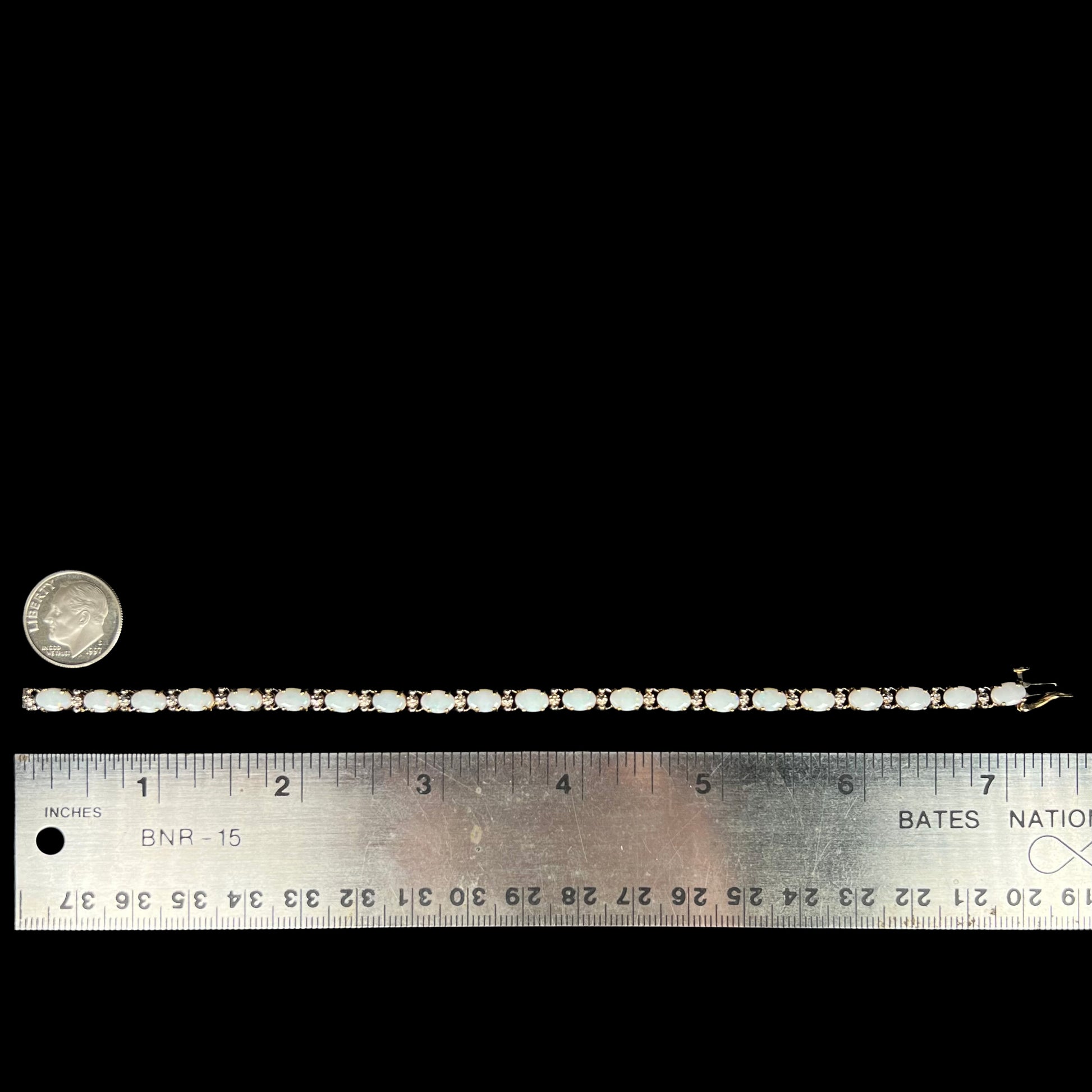 A 7 inch-long oval cut opal tennis bracelet in 10k gold shown on a black background next to a ruler and US dime coin for scale.
