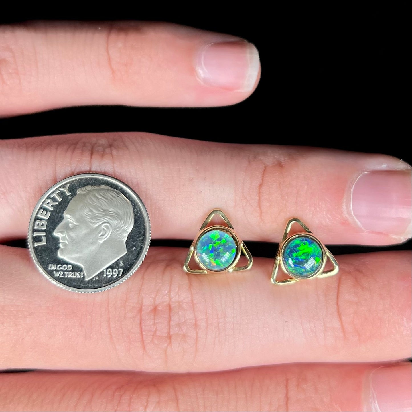 A pair of opal triangle earrings shown on top of a hand next to a 1997 S dime coin.