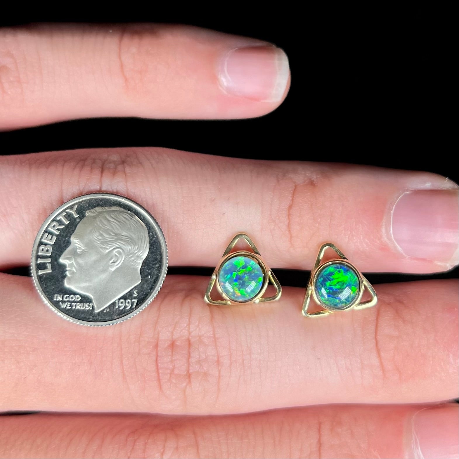 A pair of opal triangle earrings shown on top of a hand next to a 1997 S dime coin.