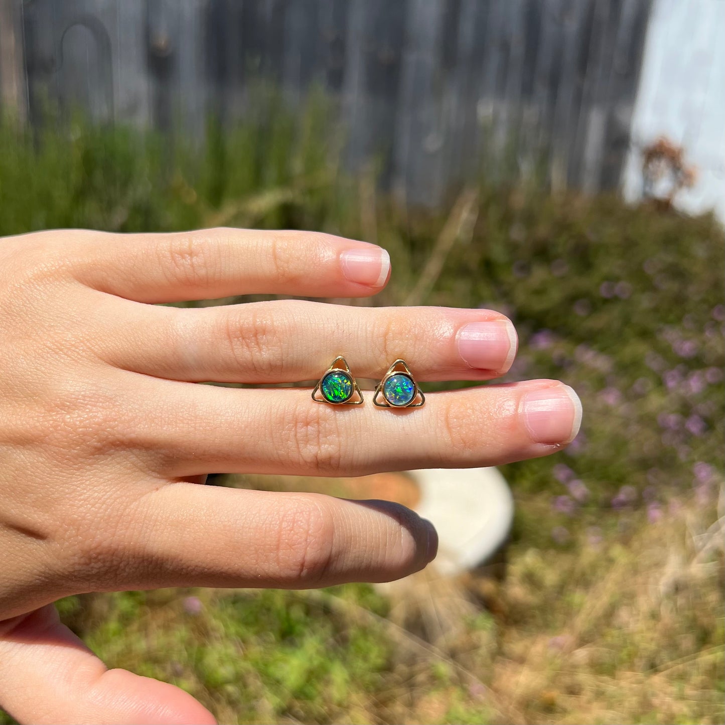 A pair of opal triangle earrings shown in the sunlight at a distance on top of a hand.  The opals appear green.