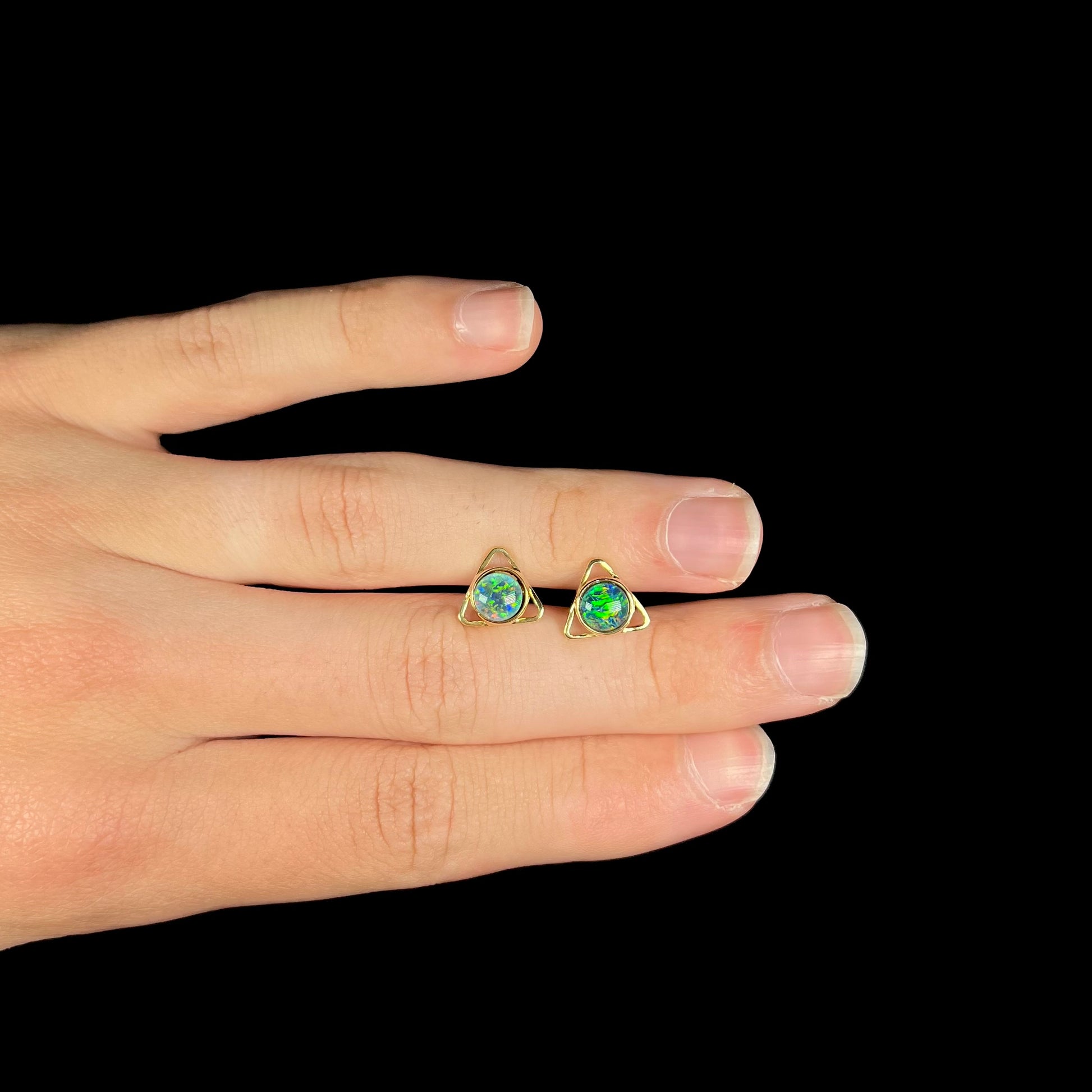 A pair of opal triplet earrings made from 18k yellow gold, held between two fingers and resting on top of a hand.