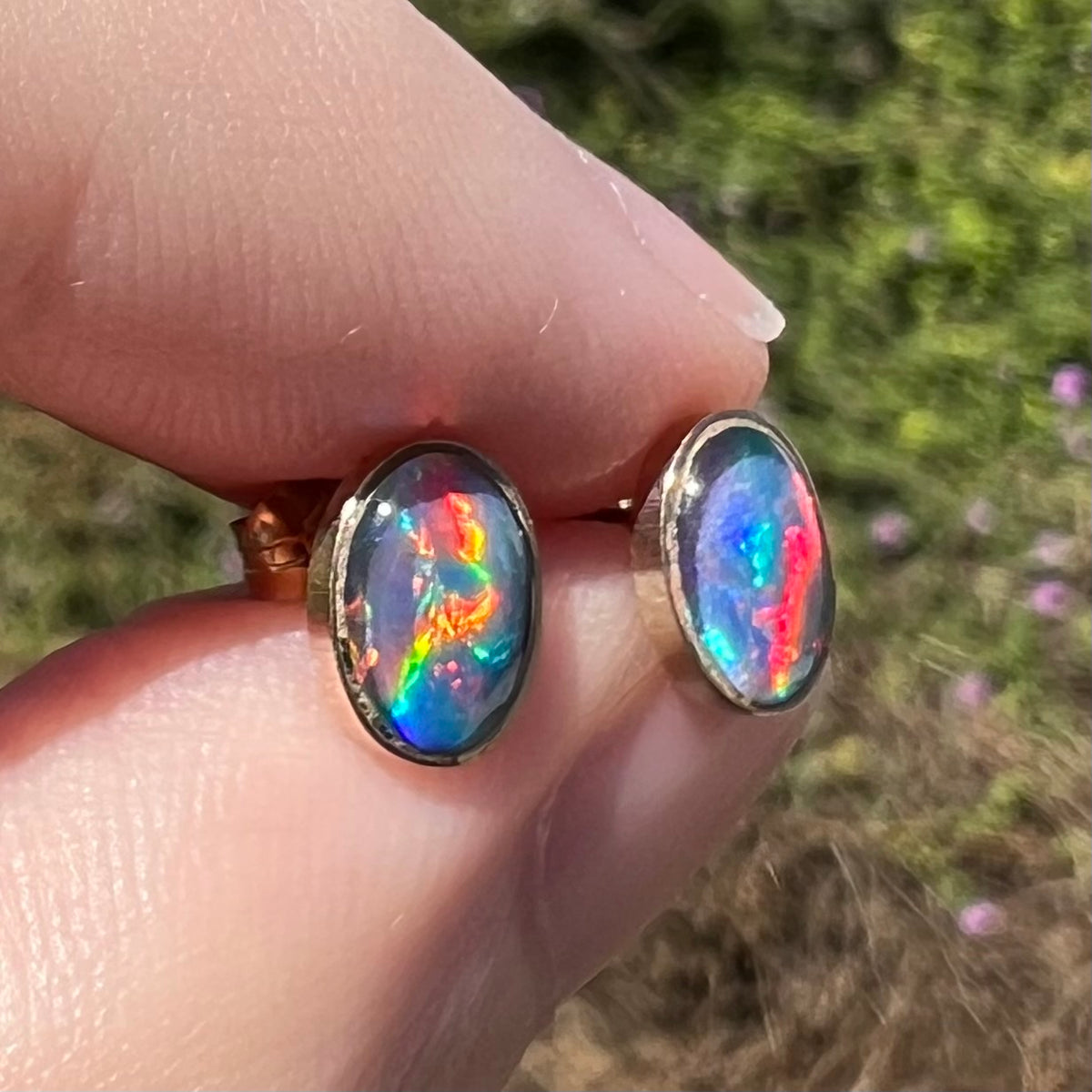 Angled close-up of opal triplet earrings in 9k gold with bright red flashes of color, held between fingers in sunlight