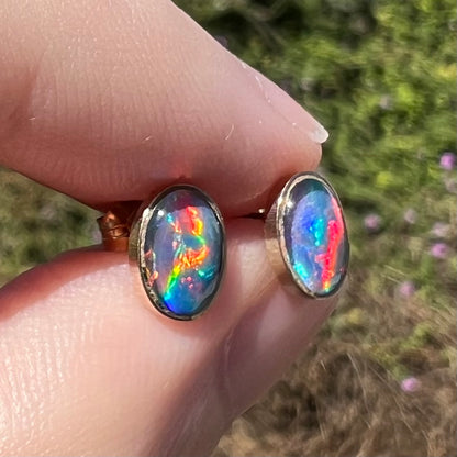 Angled close-up of opal triplet earrings in 9k gold with bright red flashes of color, held between fingers in sunlight