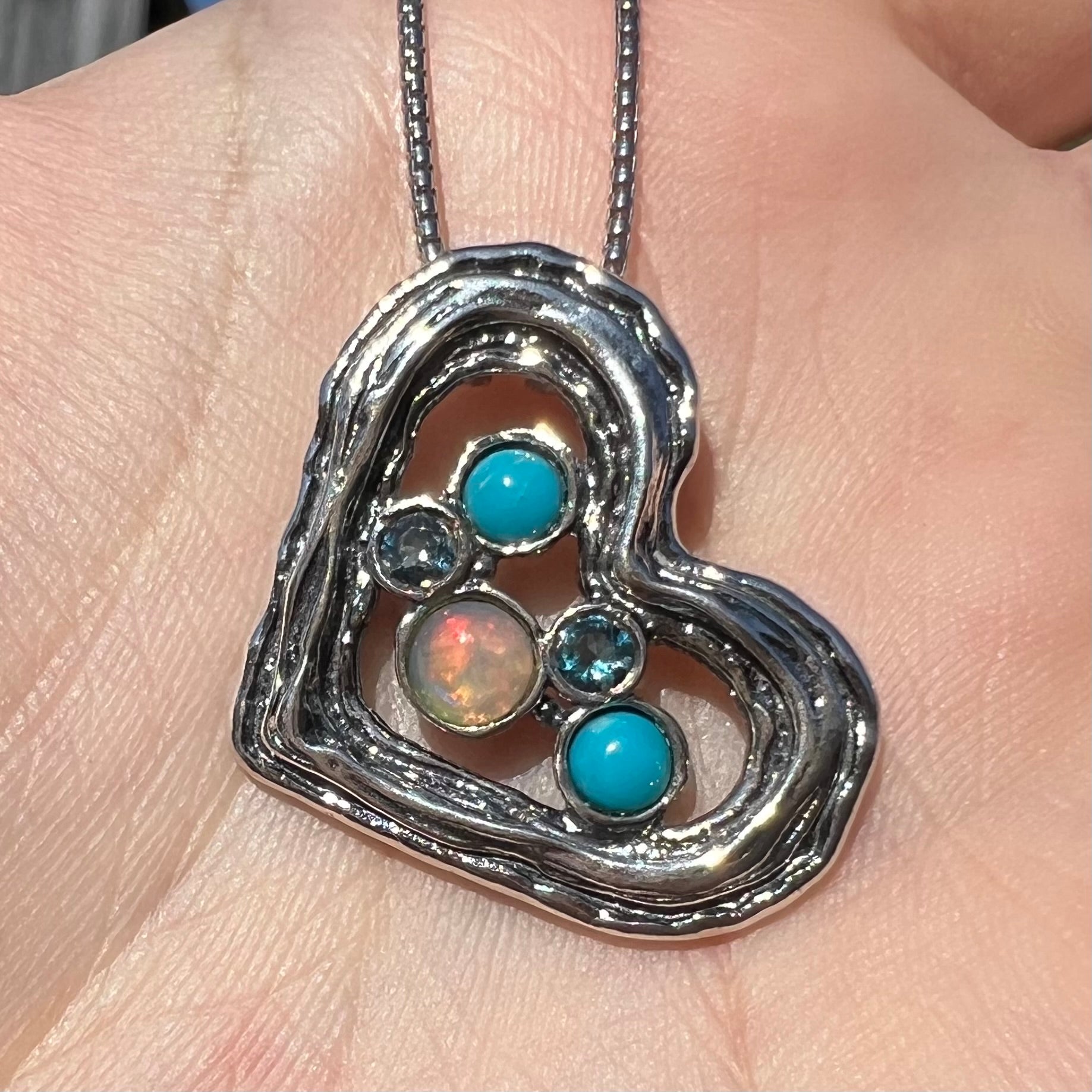 Close-up sunlit view of a sterling silver heart necklace set with opal, turquoise, and blue topaz in a hand.