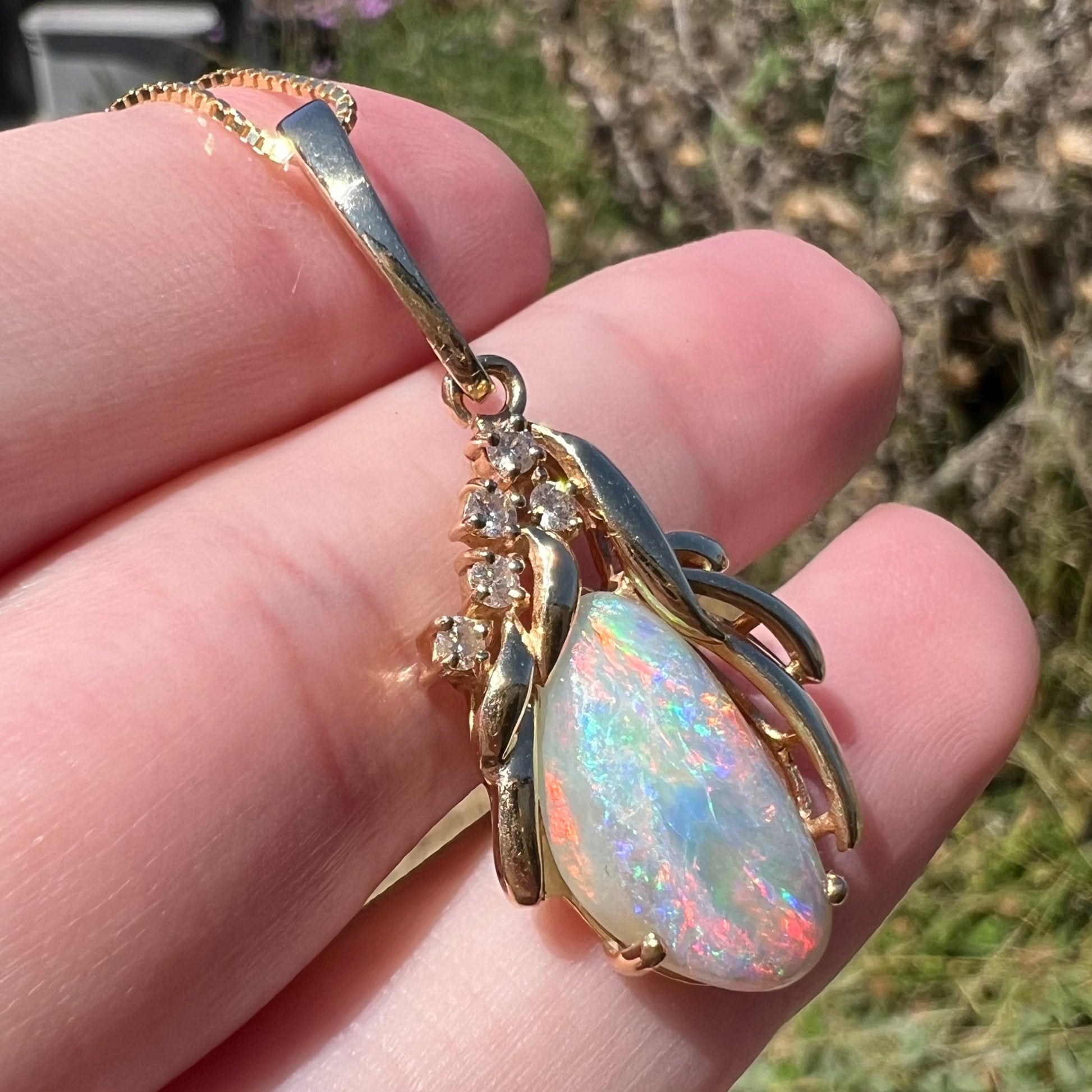 Close-up angled view of a 14k yellow gold opalized shell fossil pendant with diamond accents, held on top of fingers in sunlight.
