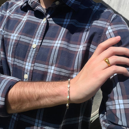 Person resting hand on shoulder while wearing blue plaid shirt, gold ring on finger, and gold opal bracelet with open link design on wrist.