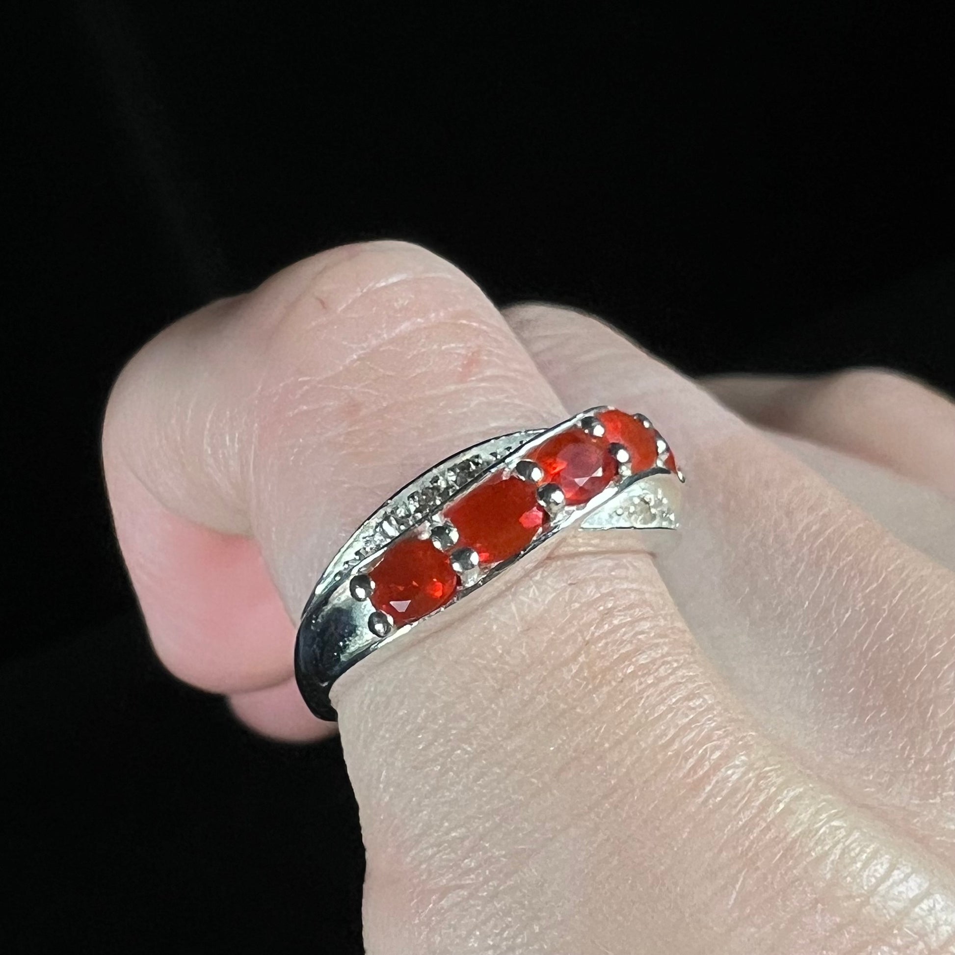 Angled close-up of silver ring with red-orange fire opal gemstones worn on a finger against black background.