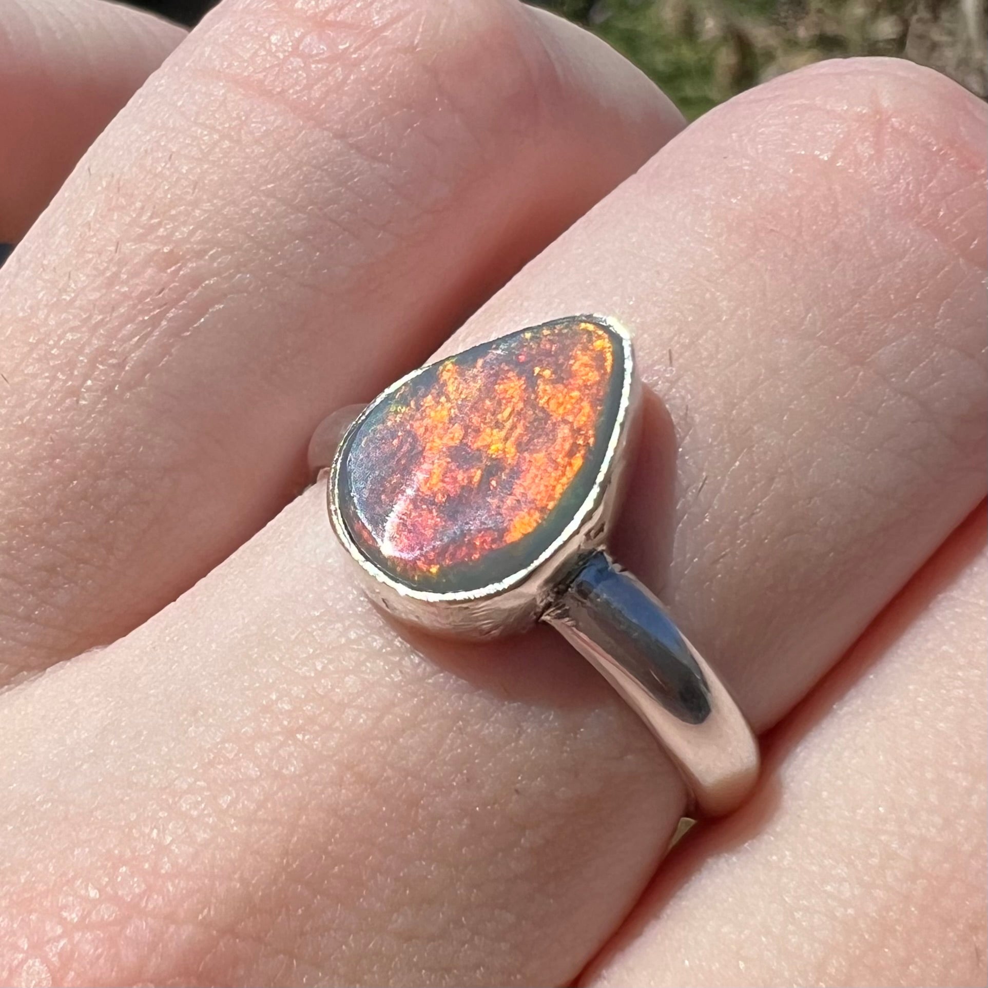 Broad orange flash of color is seen cross the surface of a pear shaped Australian dark opal ring fabricated from sterling silver, worn on a finger in sunlight.