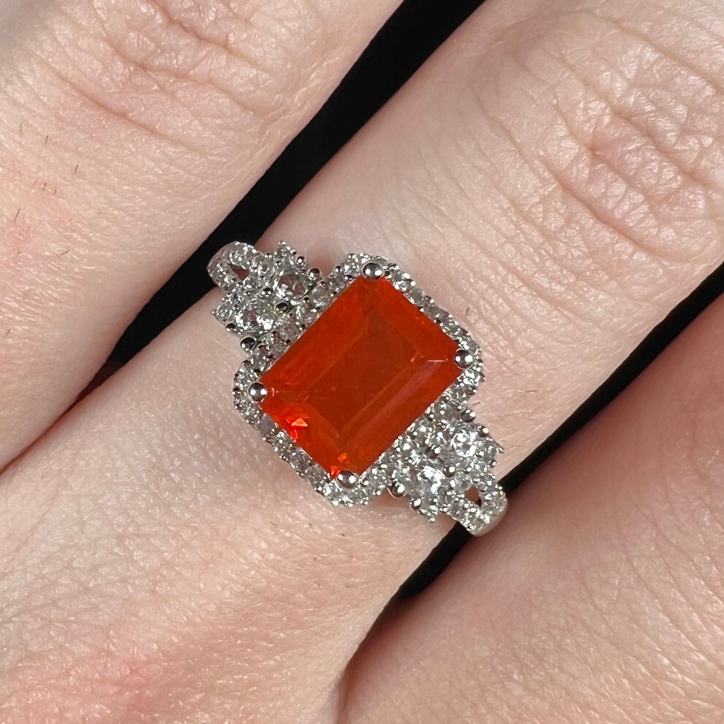 Close-up of an orange Mexican fire opal and white zircon halo ring worn on a hand under cool studio light.