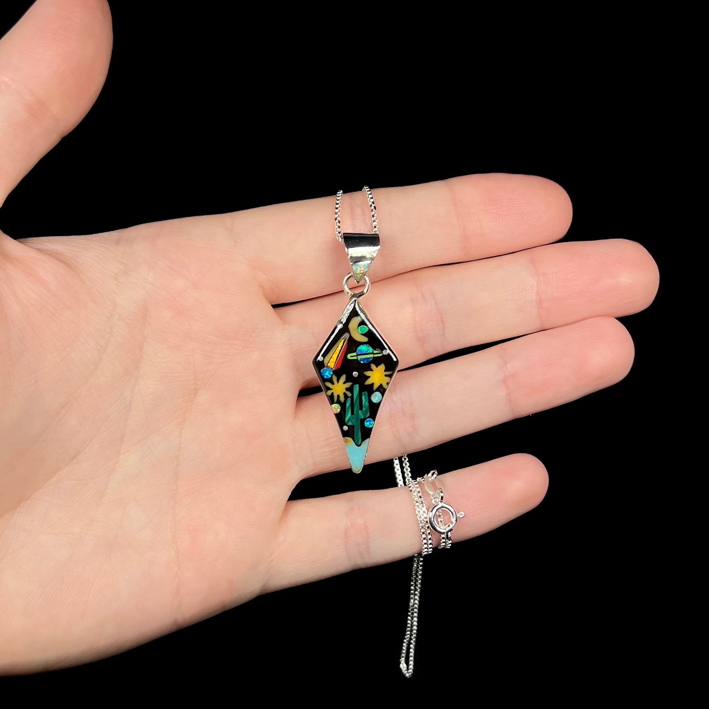 Sterling silver opal and onyx inlay necklace with outer-space motif held in hand against black background.