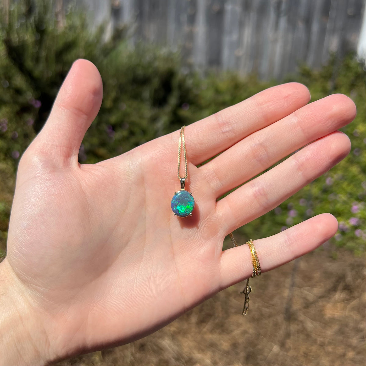 Hand holding oval shaped black opal solitaire necklace in natural sunlight; the opal has broad green flash of color