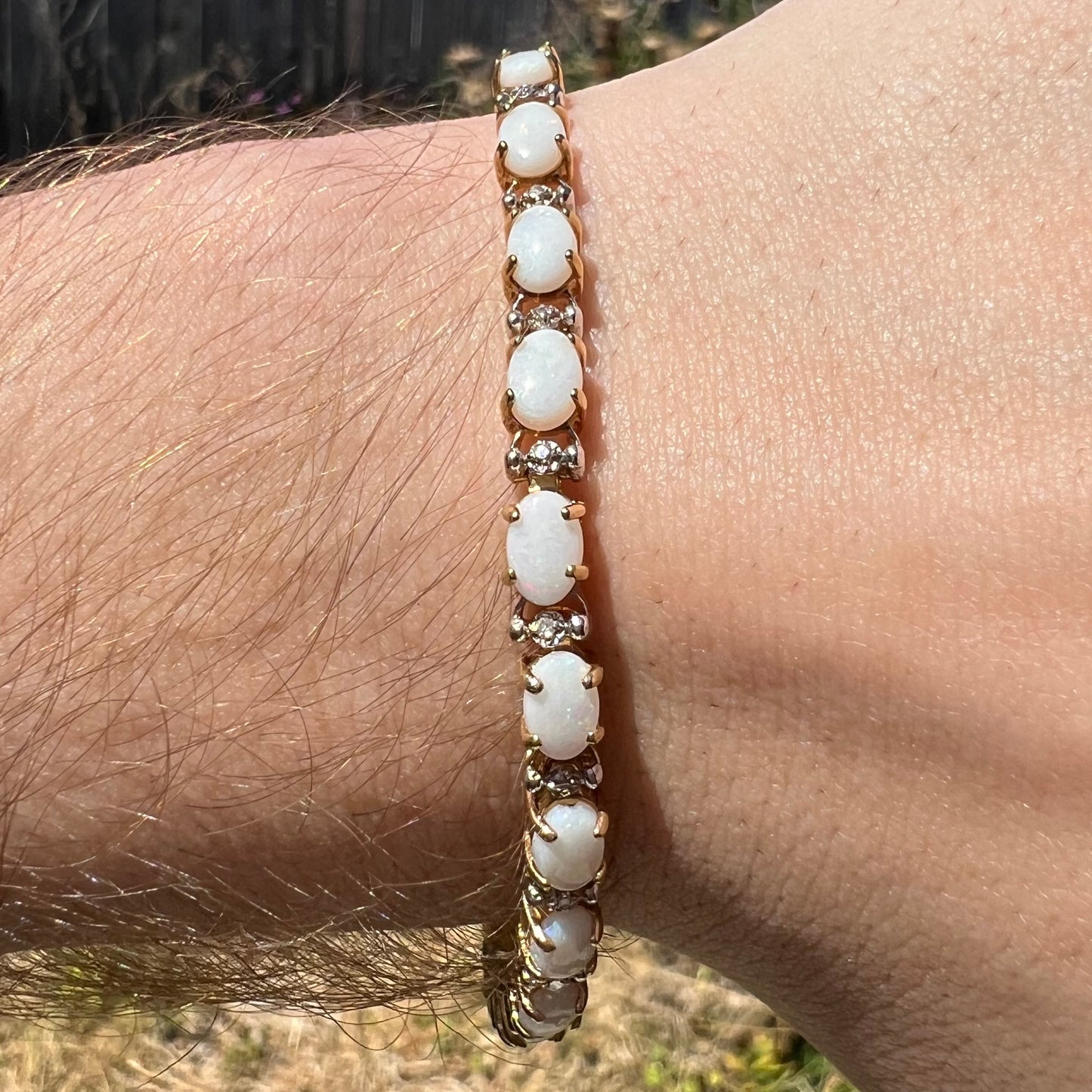 Close-up of an oval cut Australian opal and diamond tennis bracelet in gold, worn around a wrist in natural light.